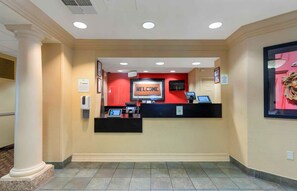 Un espace d’accueil avec un mur rouge, un panneau d’affichage indiquant « Bienvenue » et un comptoir de réception équipé d’un ordinateur et d’un téléphone.
