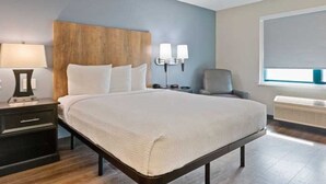 A hotel room with a large bed, a nightstand with a lamp, a wall-mounted light fixture, a chair, and a window with a view. - Extended Stay America Suites Oakland Alameda Airport (Oakland)