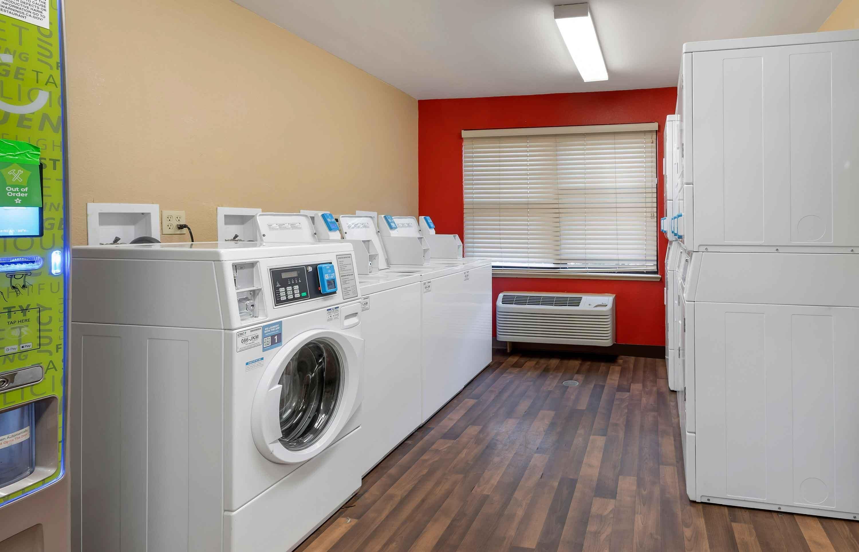 A laundry room with multiple washing machines, a dryer, and a window with blinds.