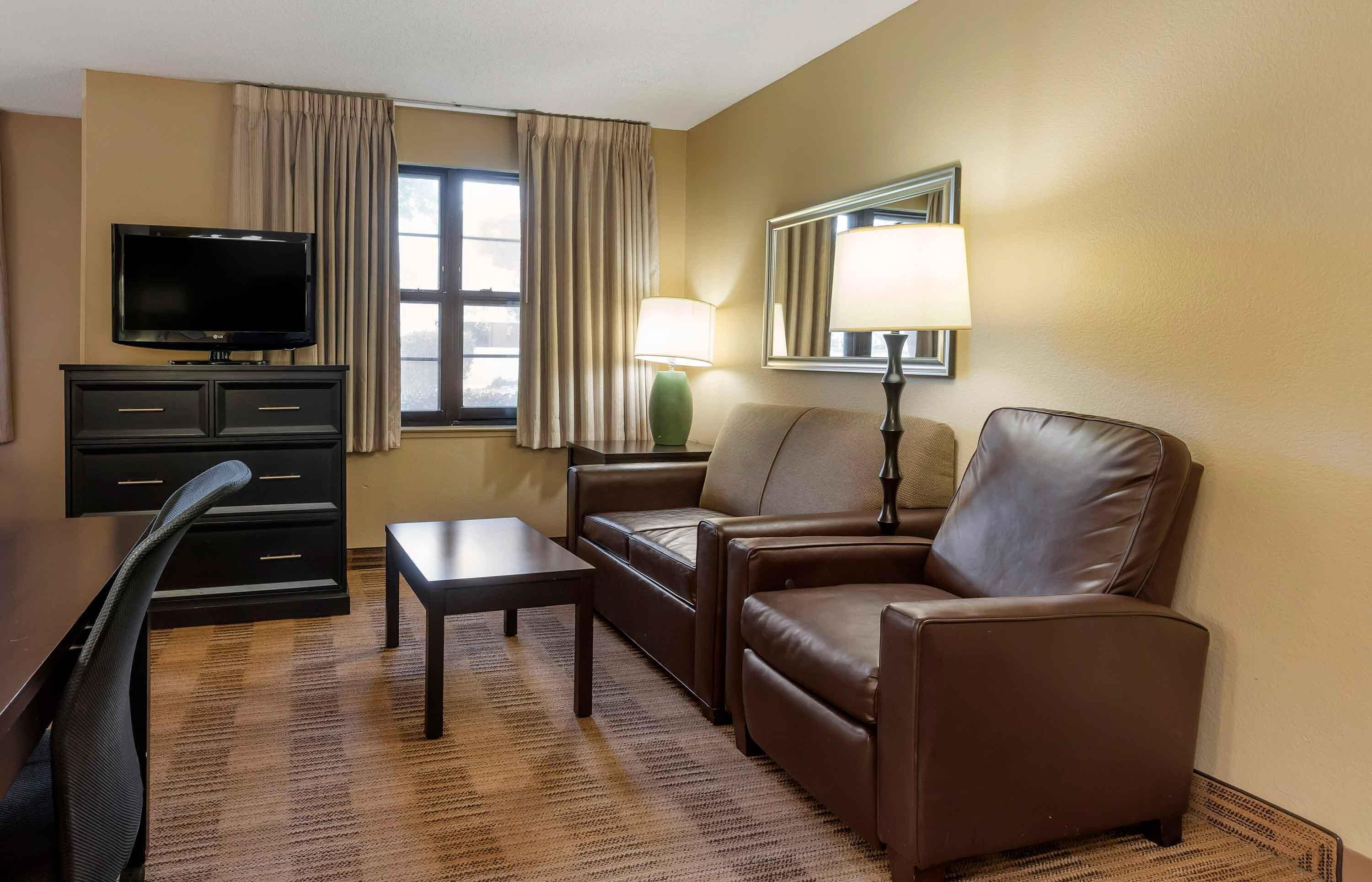 Habitación de hotel con televisión de pantalla plana, sofá, mesa de centro, lámpara y ventana con cortinas.
