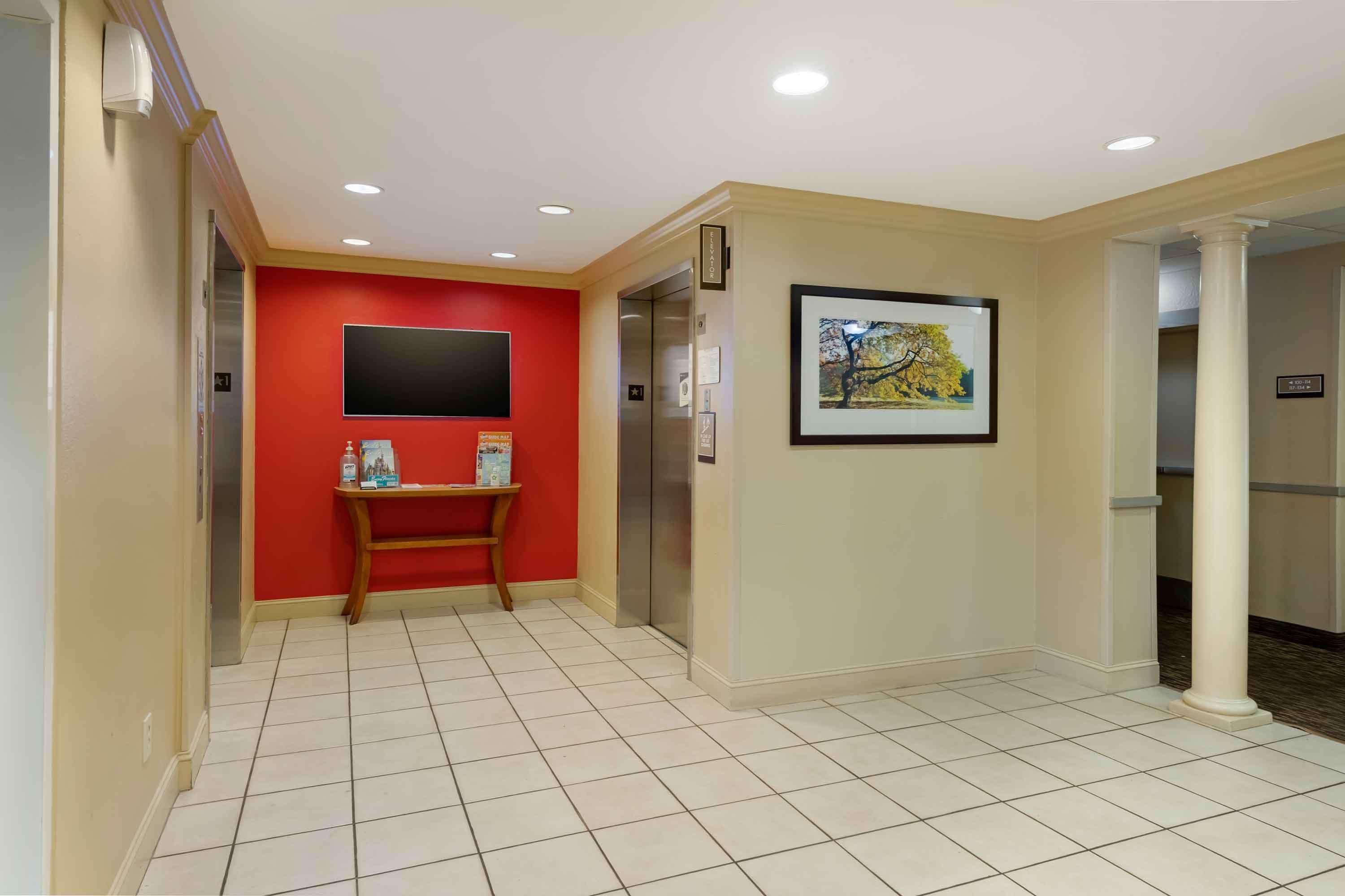 Een hotellobby met een rode accentmuur, een wandgemonteerde televisie, een kunstwerk in een lijst en een betegelde vloer.