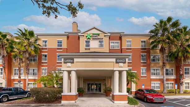 Un hotel Stay America con entrada al vestíbulo, palmeras y zona de estacionamiento.
