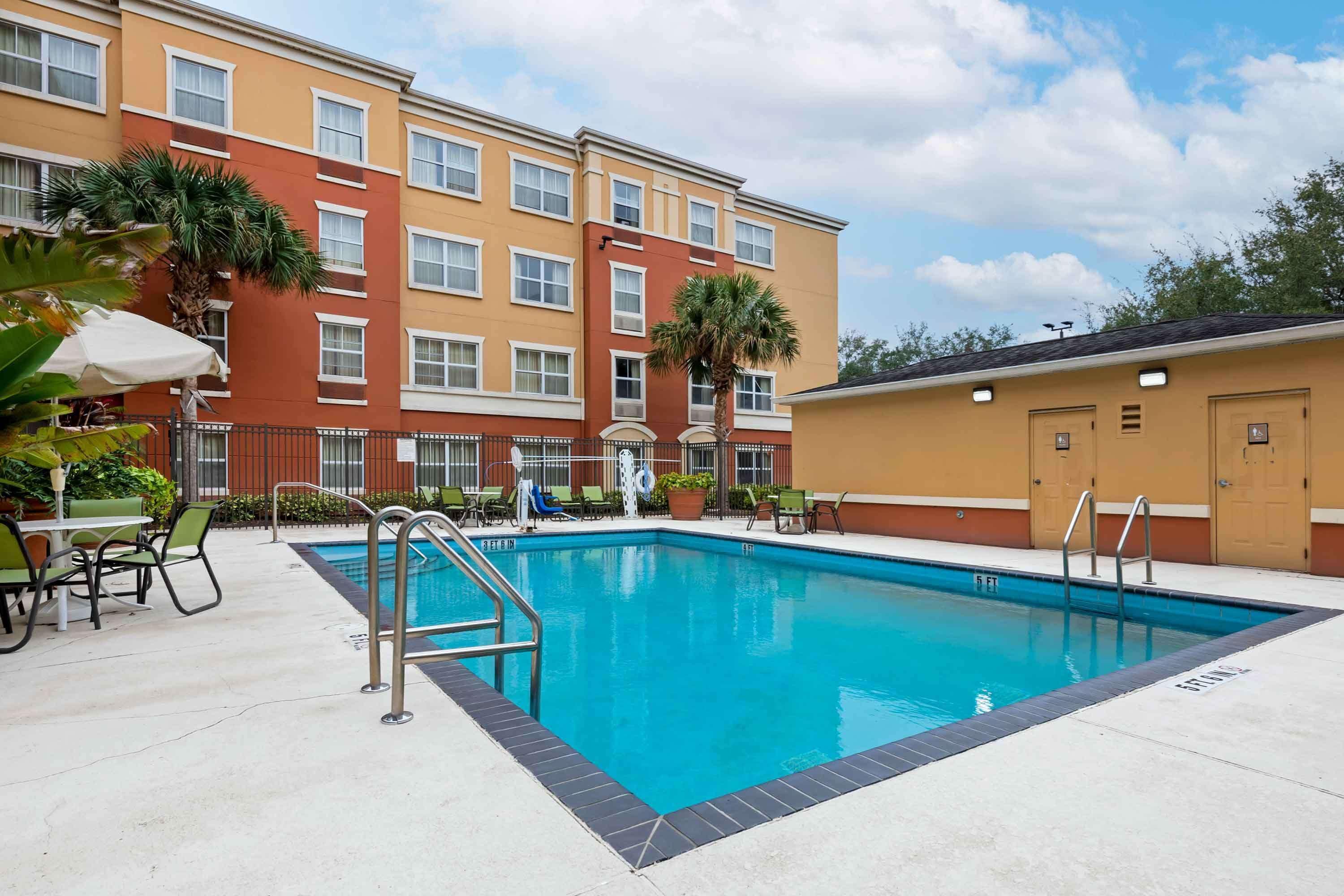 A swimming pool with a metal ladder, surrounded by a patio area with chairs and tables, and a multi-story building with a red and beige facade in the background.