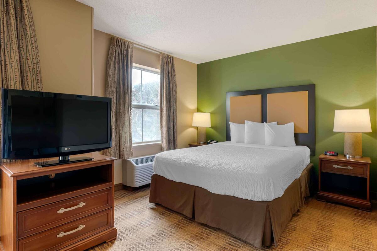 A hotel room with a bed, a TV, a nightstand, and a window.