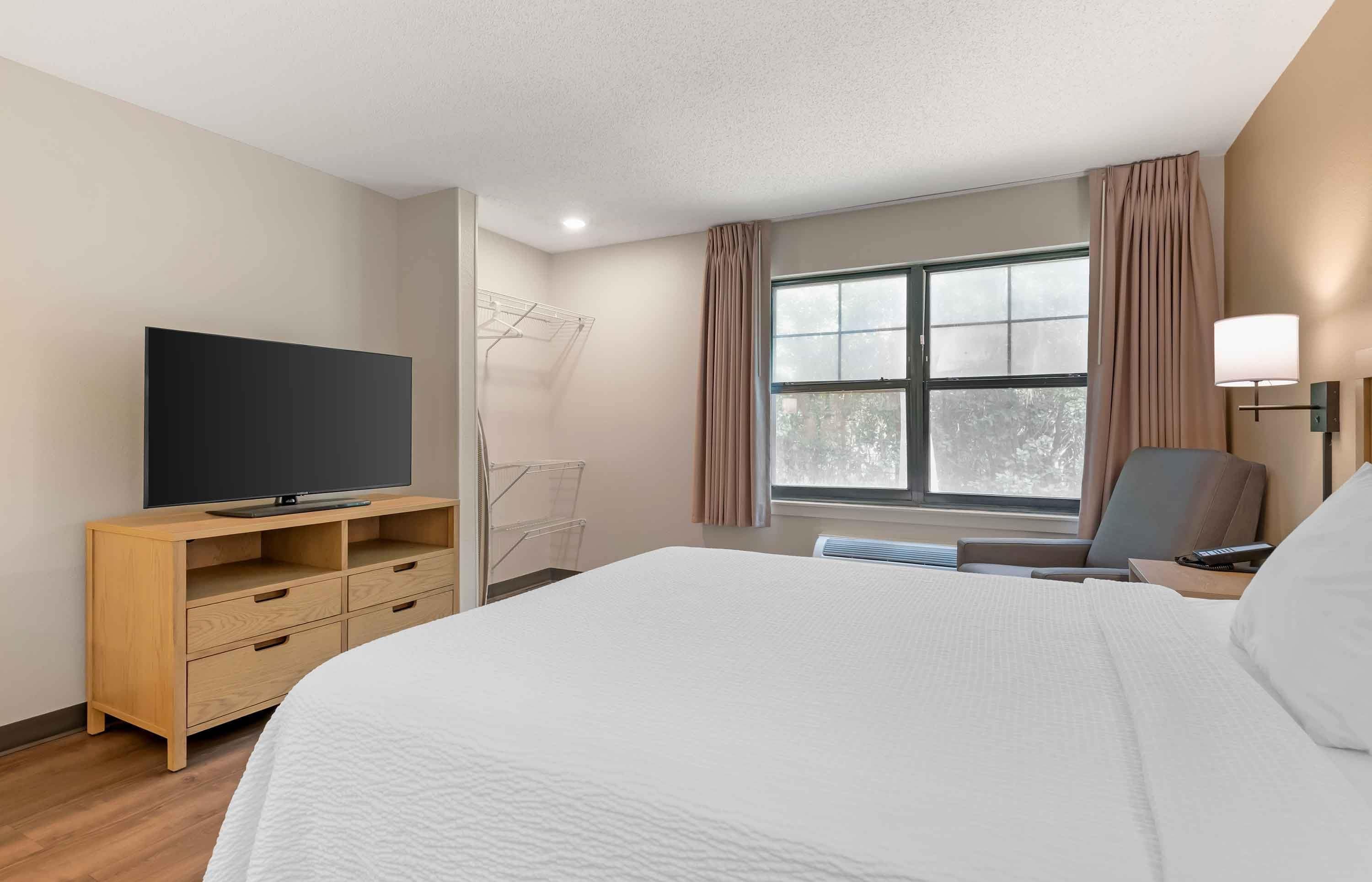 Habitación de hotel con cama, televisión sobre un soporte de madera, una silla, un escritorio con lámpara y una ventana con cortinas.