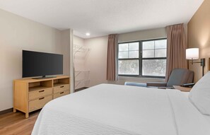 Een hotelkamer met een bed, een televisie op een houten standaard, een stoel, een bureau met een lamp, en een raam met gordijnen.