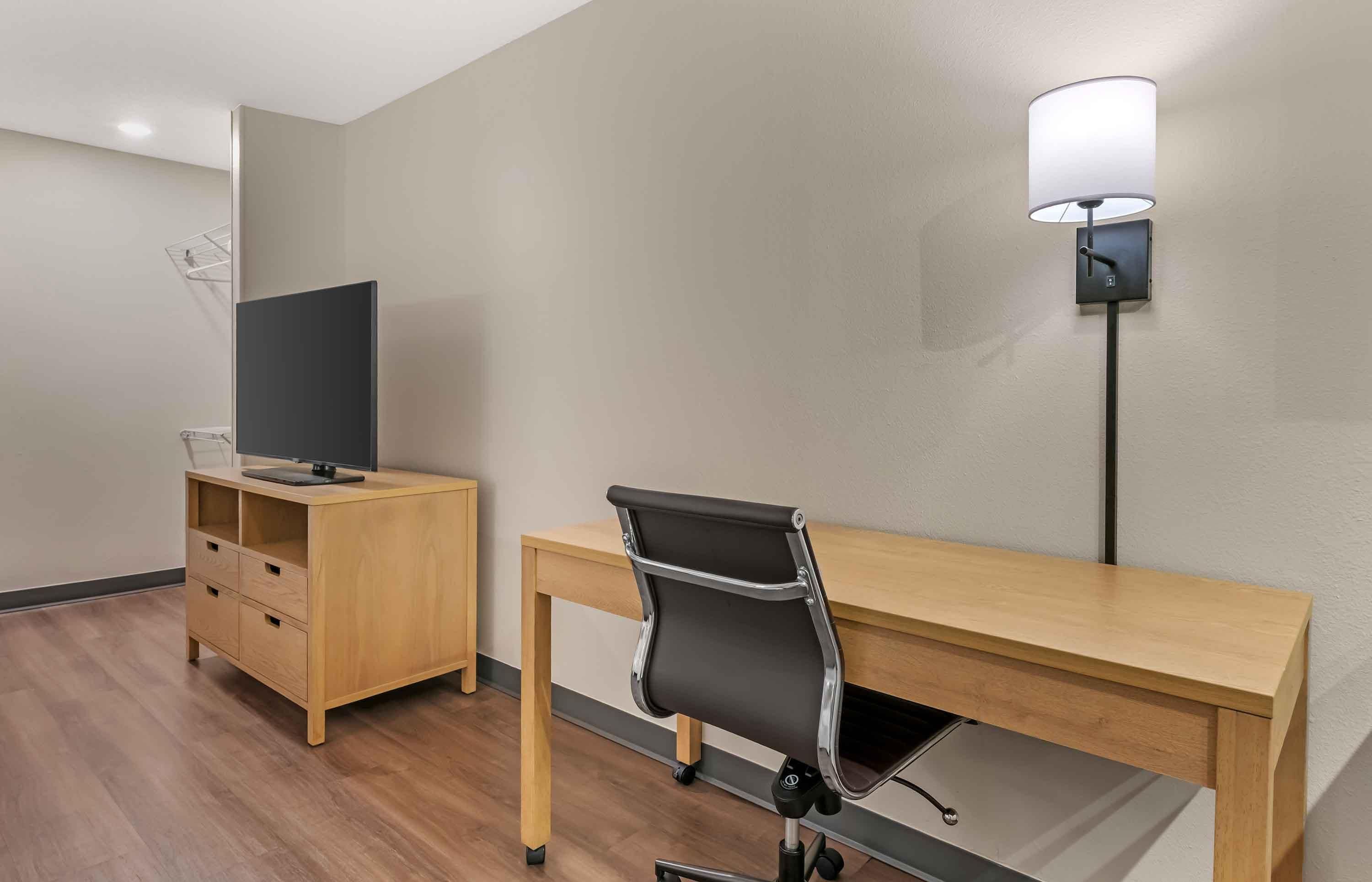 Una habitación de hotel con un escritorio de madera, una silla de oficina negra, un televisor sobre un mueble de madera y una lámpara fijada a la pared.