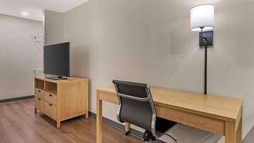 Una habitación de hotel con un escritorio de madera, una silla de oficina negra, un televisor sobre un mueble de madera y una lámpara fijada a la pared.