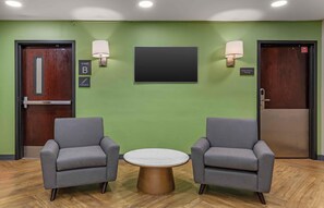 Een hal met twee grijze fauteuils, een ronde tafel, een flatscreen-televisie en twee deuren met de aanduidingen 'STAIR B' en 'ELECTRICAL ROOM'.