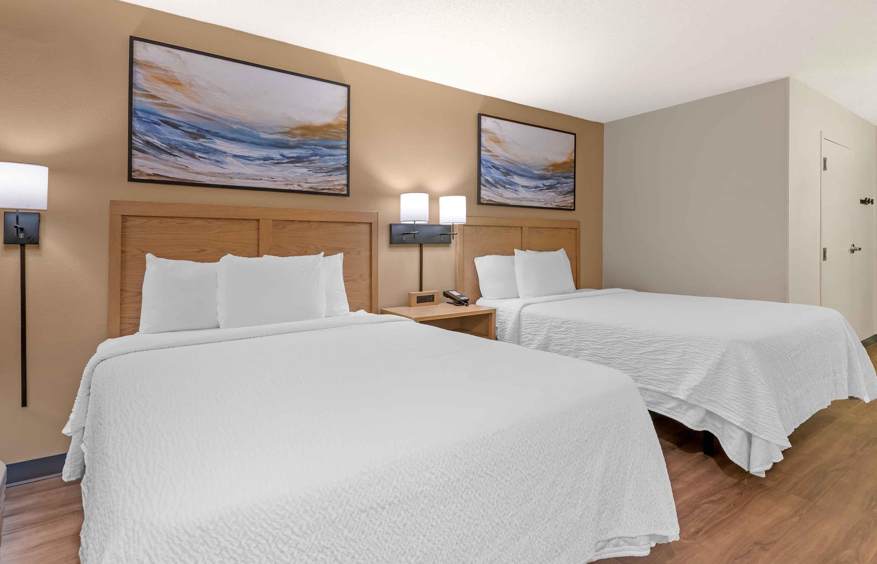 A hotel room with two beds, a nightstand with a phone, wall-mounted lamps, and two paintings.
