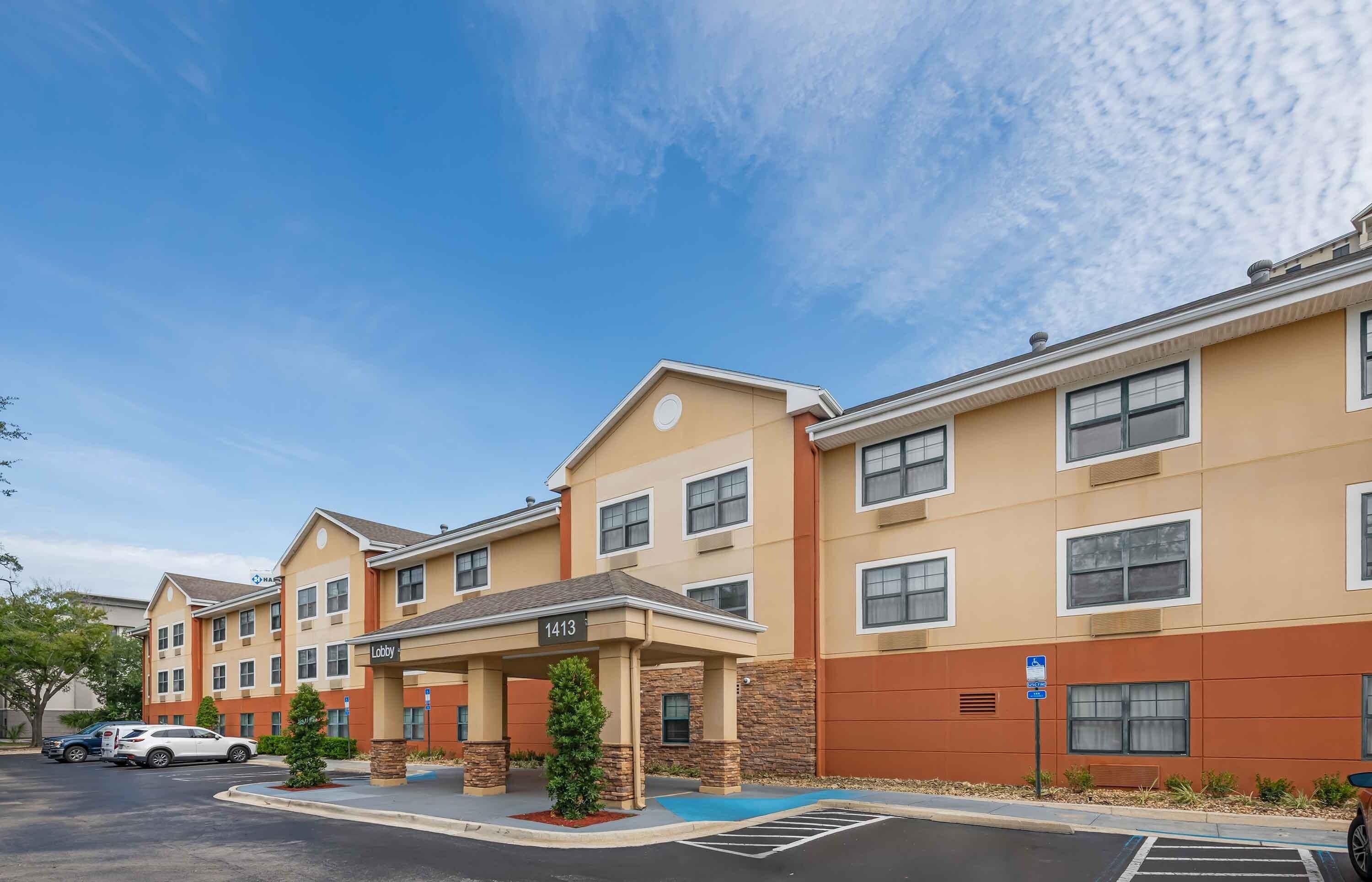 A multi-story hotel with a covered lobby area, parking lot, and a clear blue sky.