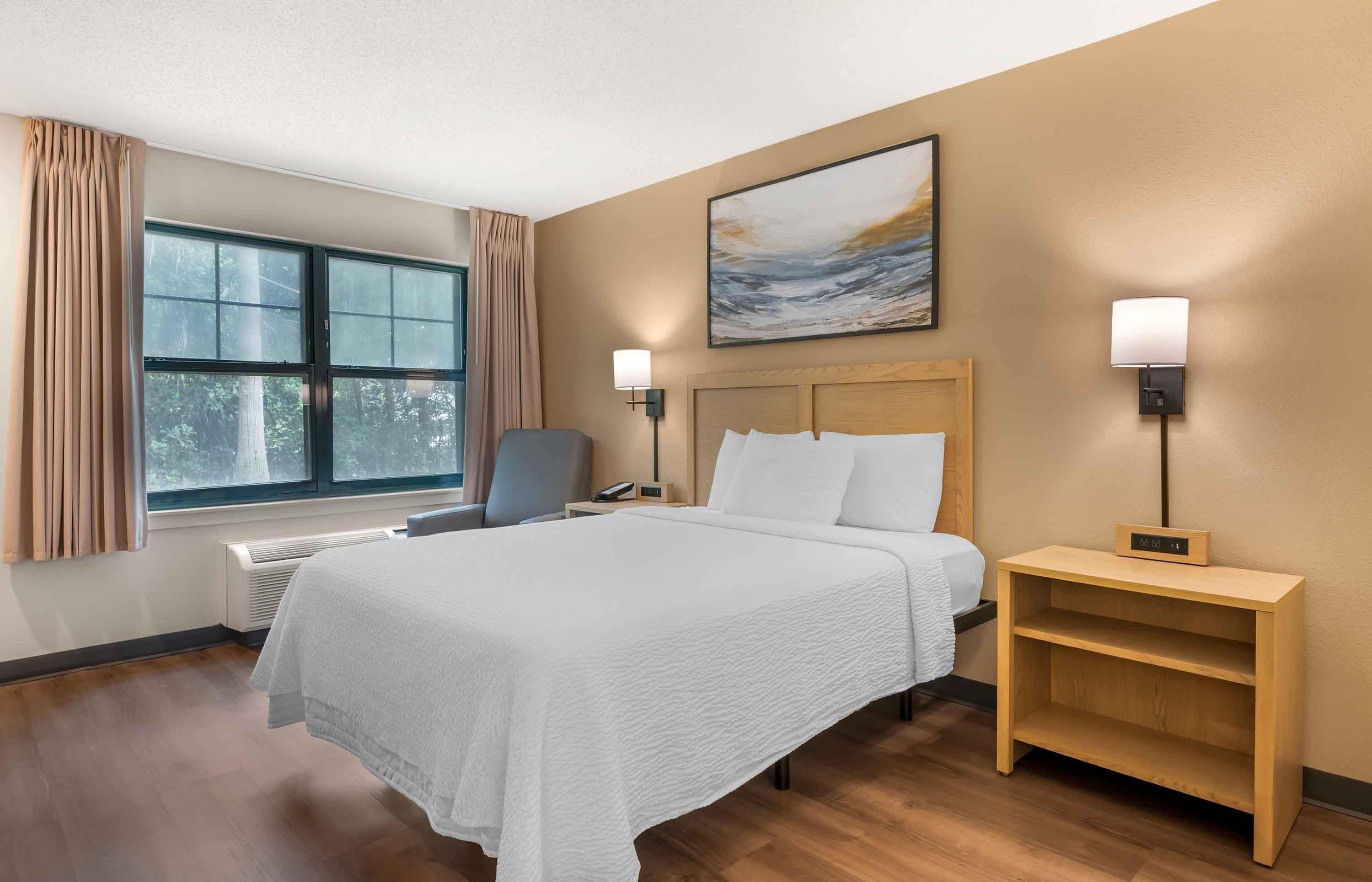 A hotel room with a bed, bedside tables, a chair, a window with curtains, and a painting on the wall.