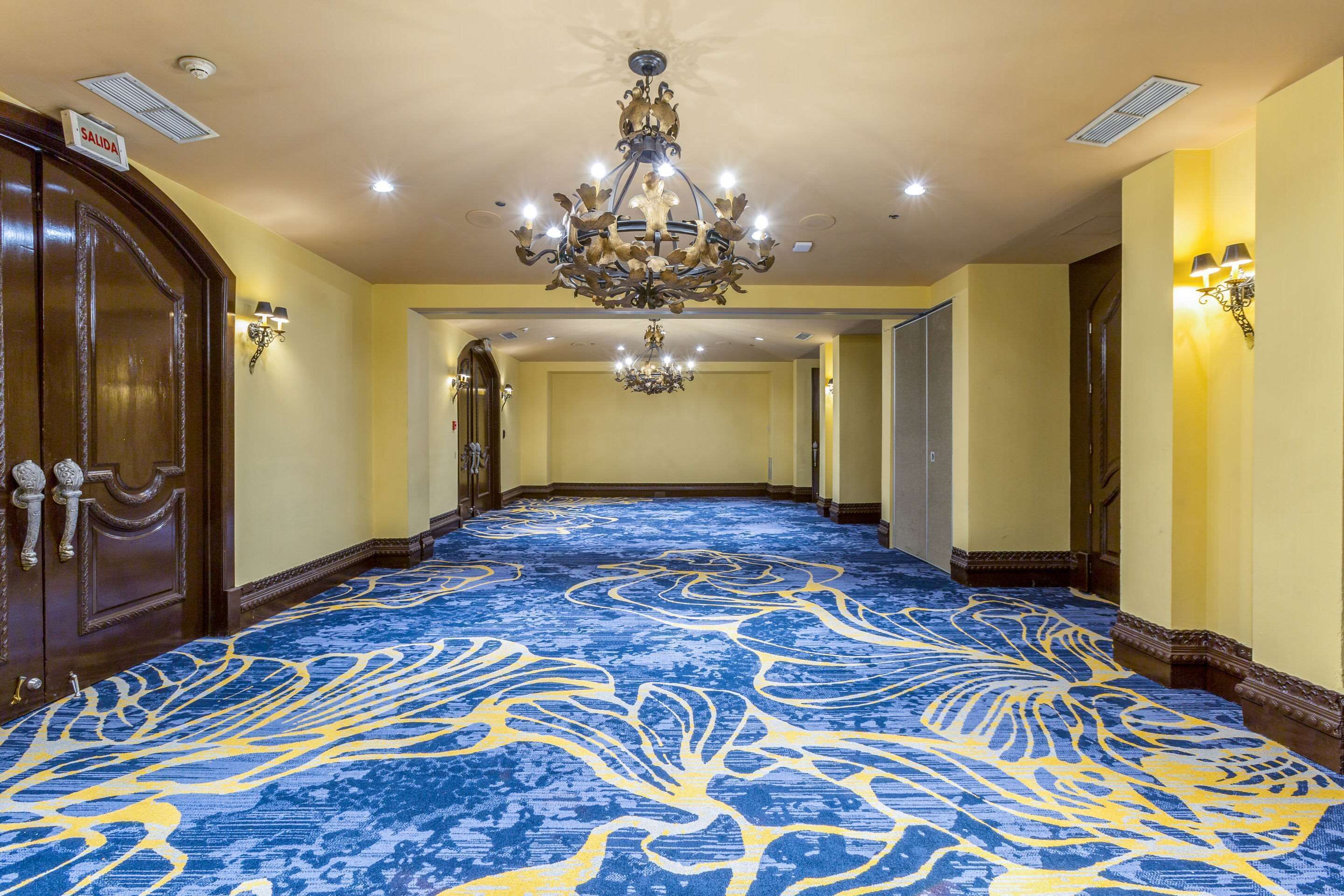Un corridoio d'hotel con tappeto blu, lampadari e porte in legno.