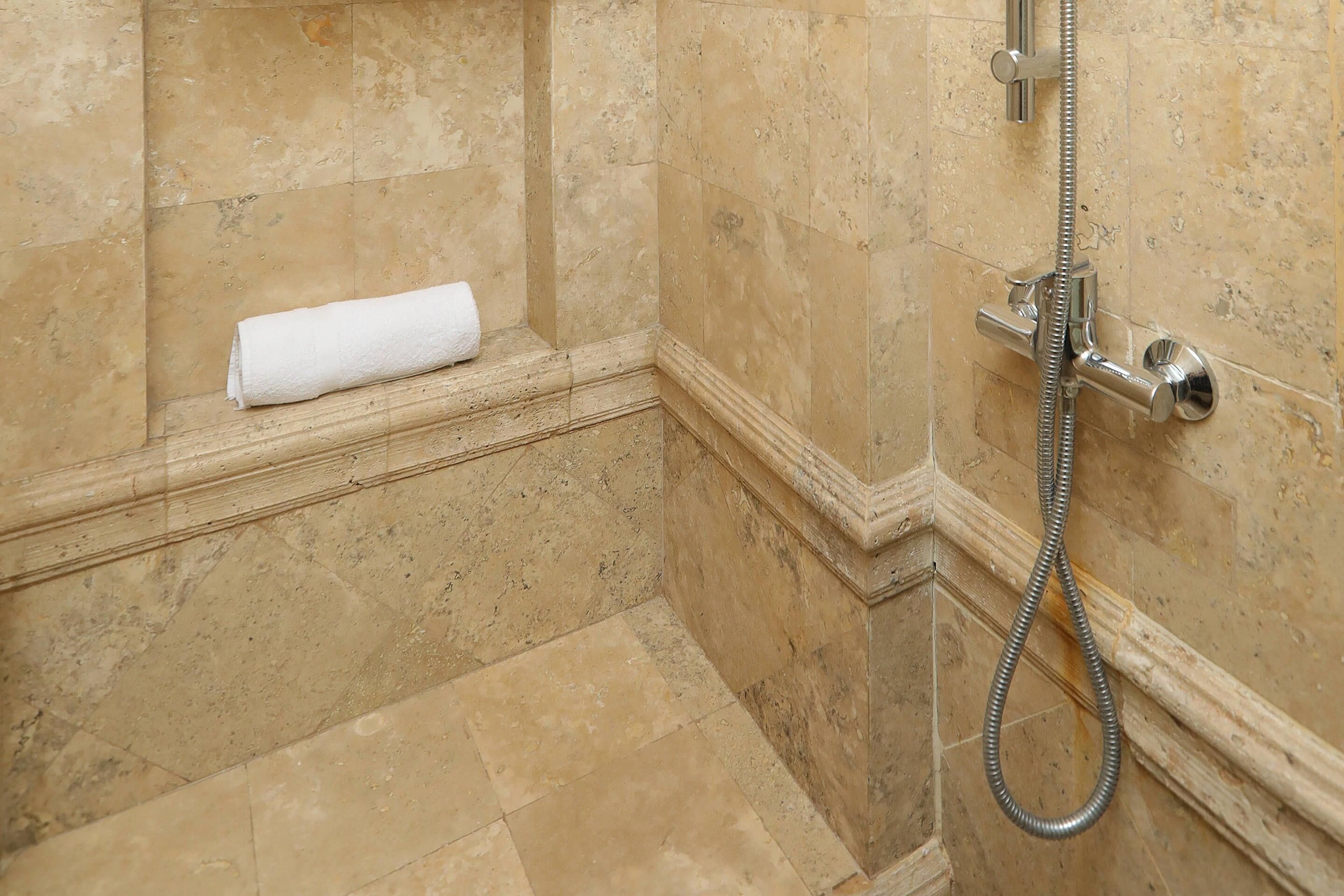 A bathroom with beige tiled walls and floor, a white towel on a shelf, and a shower with a handheld hose.