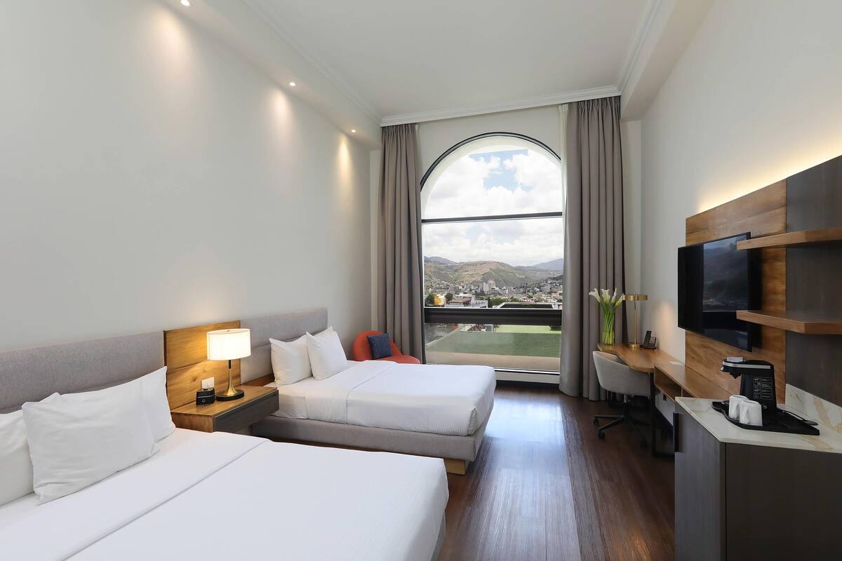 Habitación de hotel con dos camas, un escritorio, televisor y un ventanal con vistas a una ciudad y montañas.