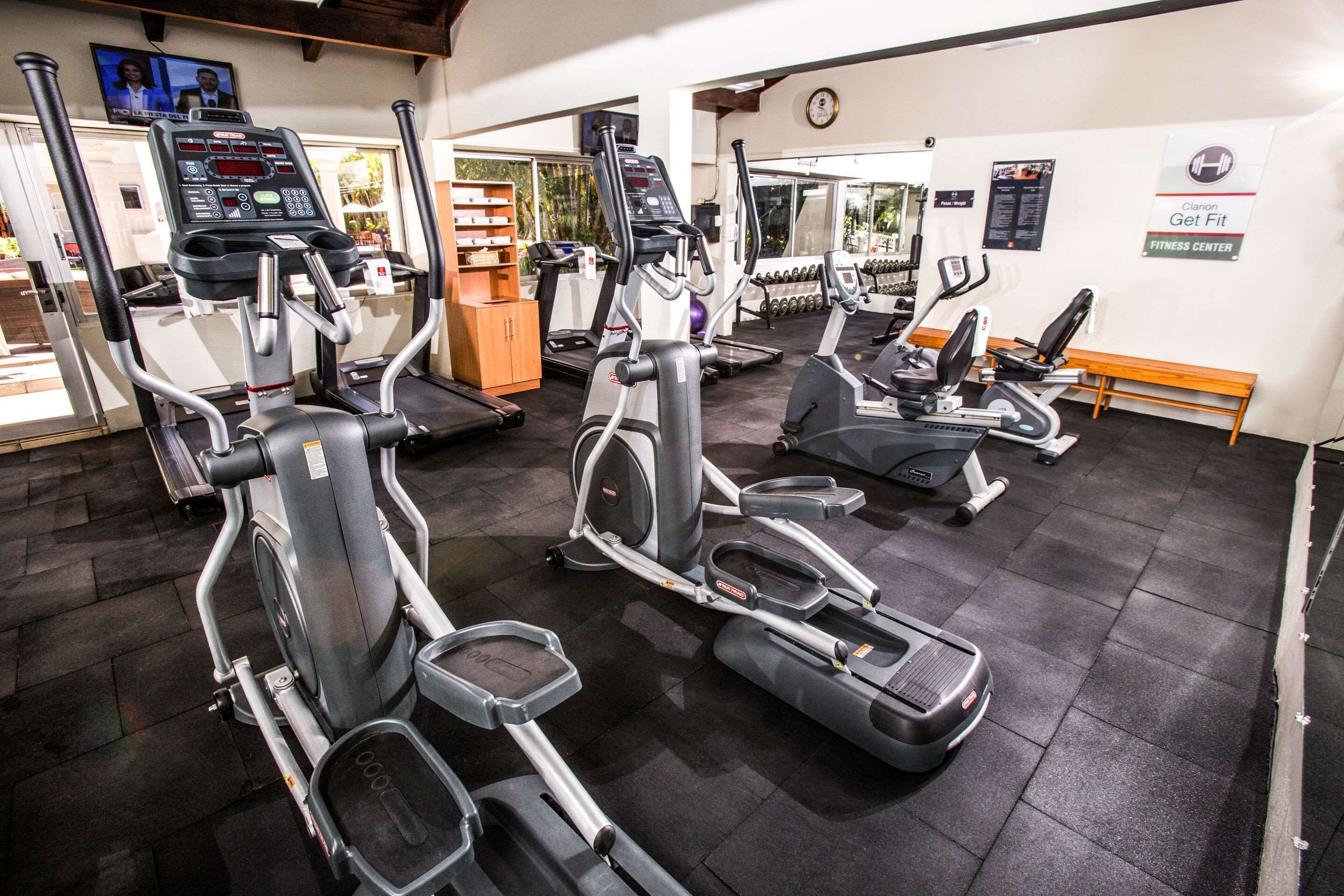 A gym with multiple cardio machines, a weight bench, and a mirror reflecting the equipment.