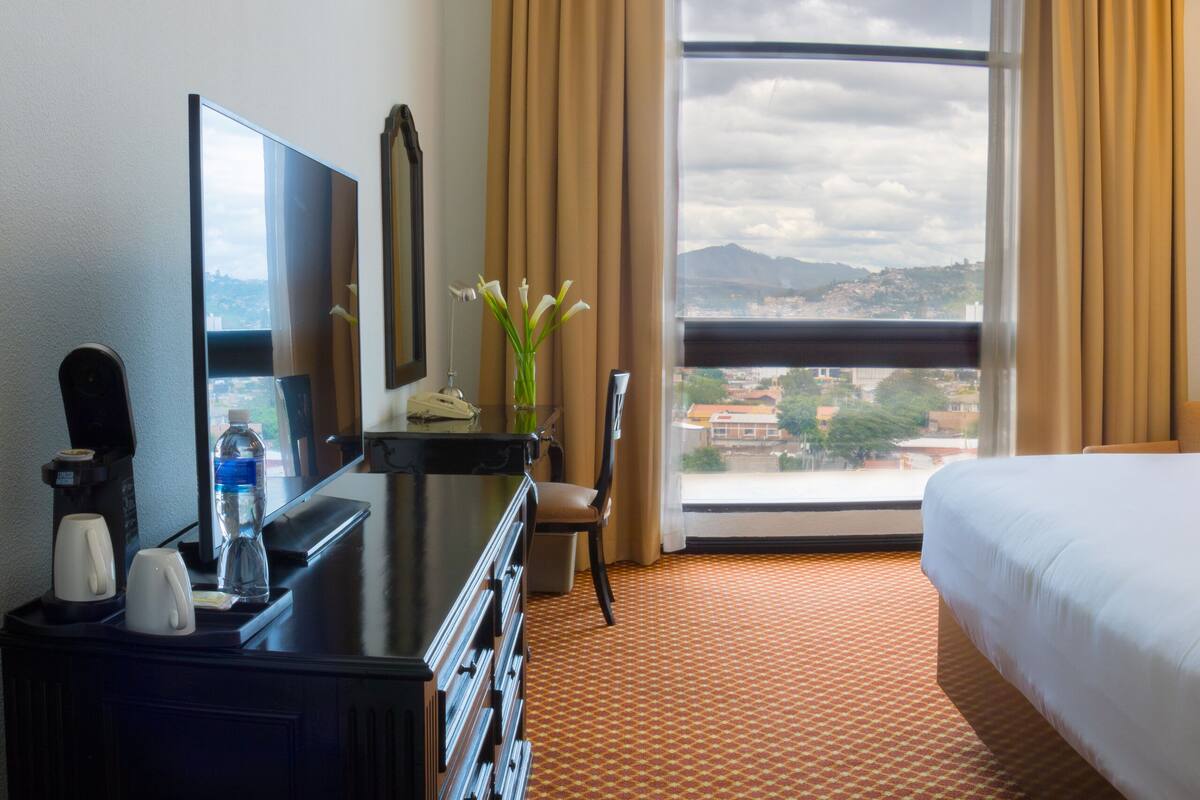 Habitación de hotel con cama, escritorio con cafetera, silla, ventana con vista a la ciudad y lámpara.