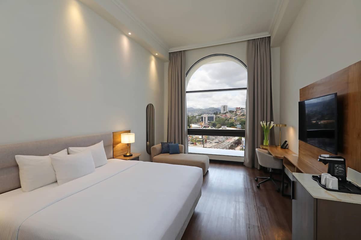Habitación de hotel con una cama grande, un escritorio, una silla, un televisor y vistas a la ciudad a través de un amplio ventanal.