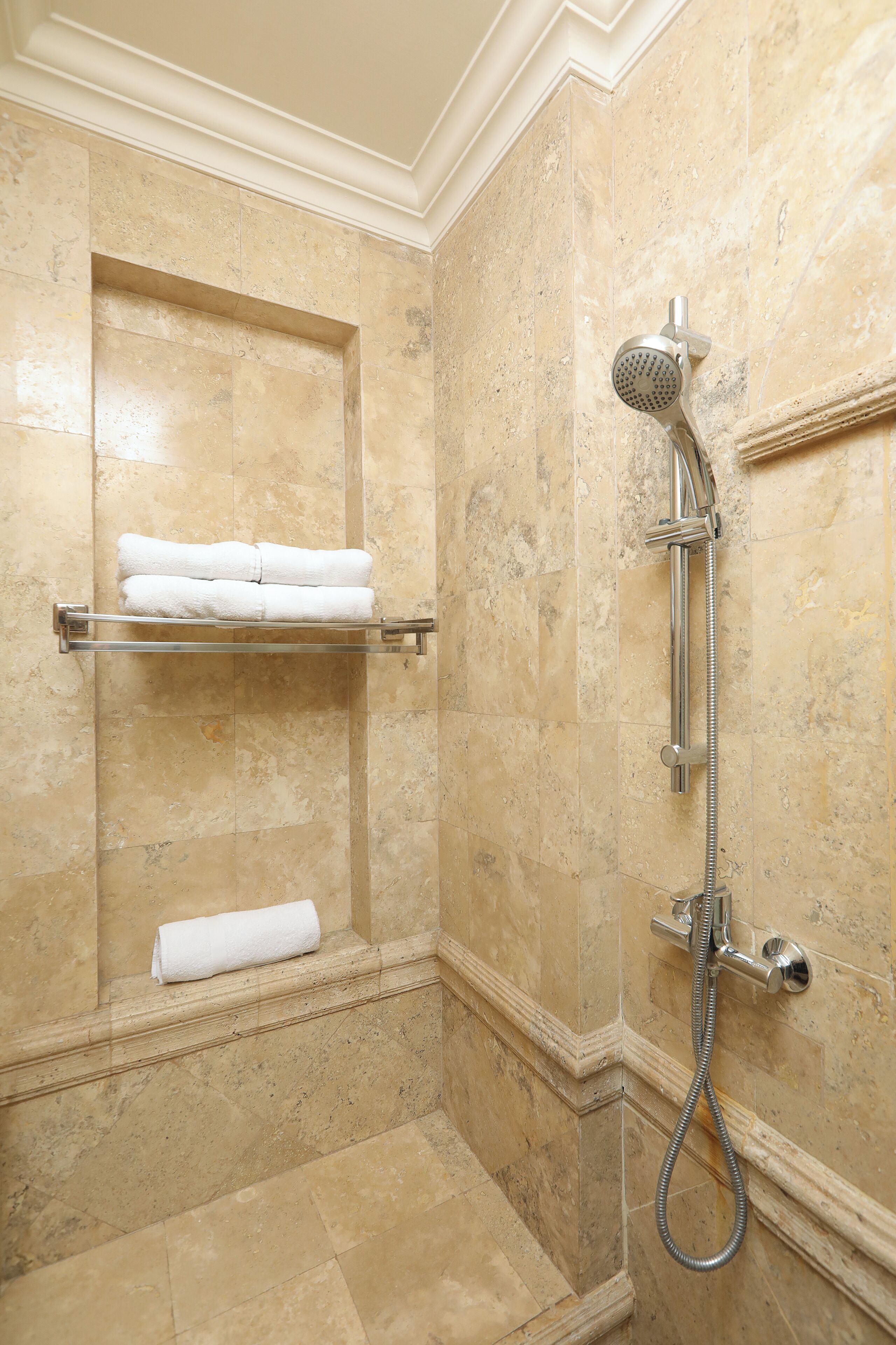 Ein Badezimmer mit beigen Marmorfliesen, einer Dusche inklusive Handbrause und einem Handtuchhalter mit gefalteten Handtüchern.