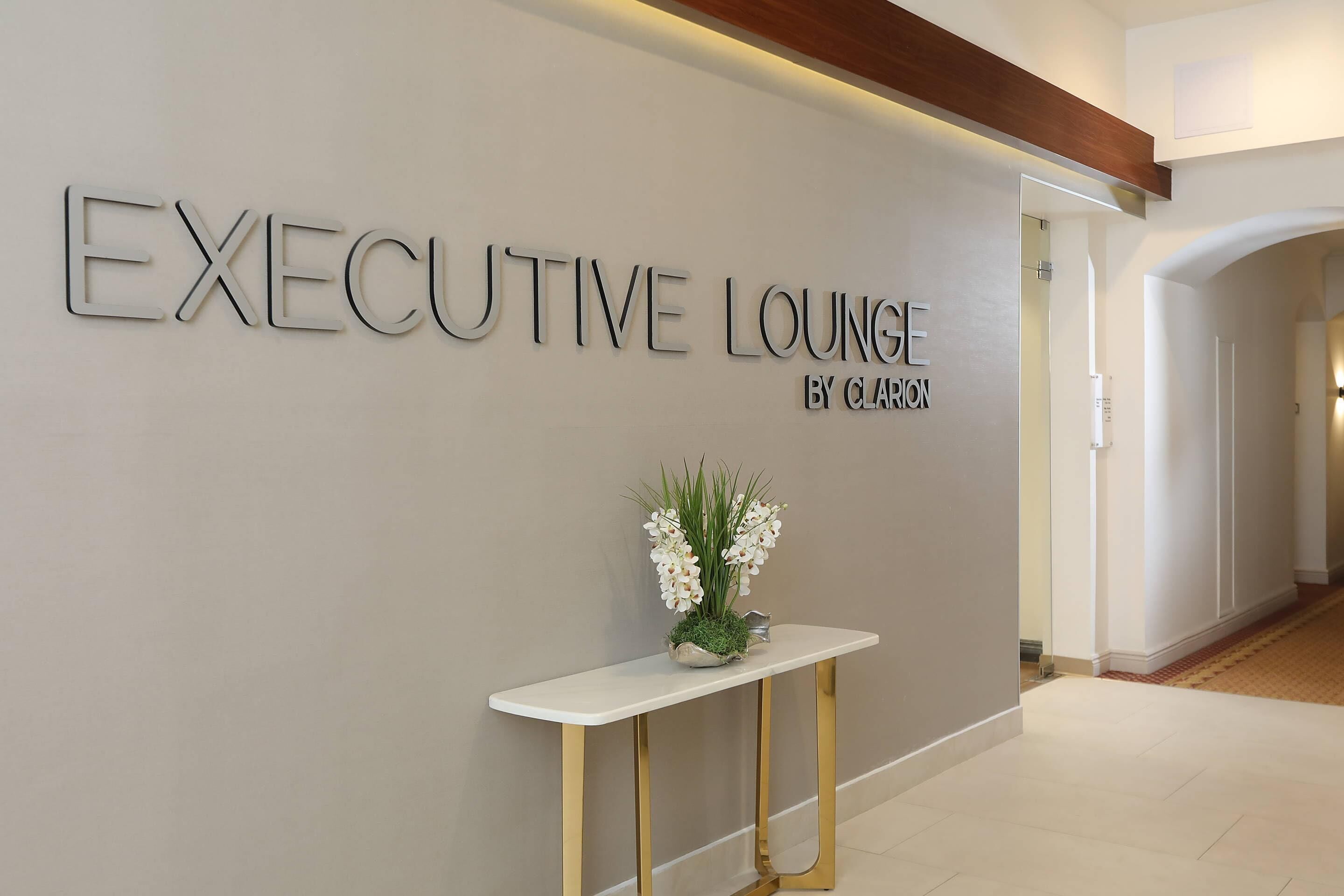Un área de recepción con un letrero que dice “EXECUTIVE LOUNGE BY CLARION” y una pequeña mesa con una planta.
