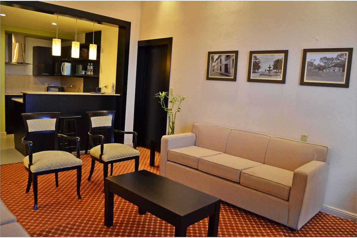 Una sala de estar con sofá, dos sillones, una mesa de centro y una cocina con refrigerador, microondas e iluminación colgante.