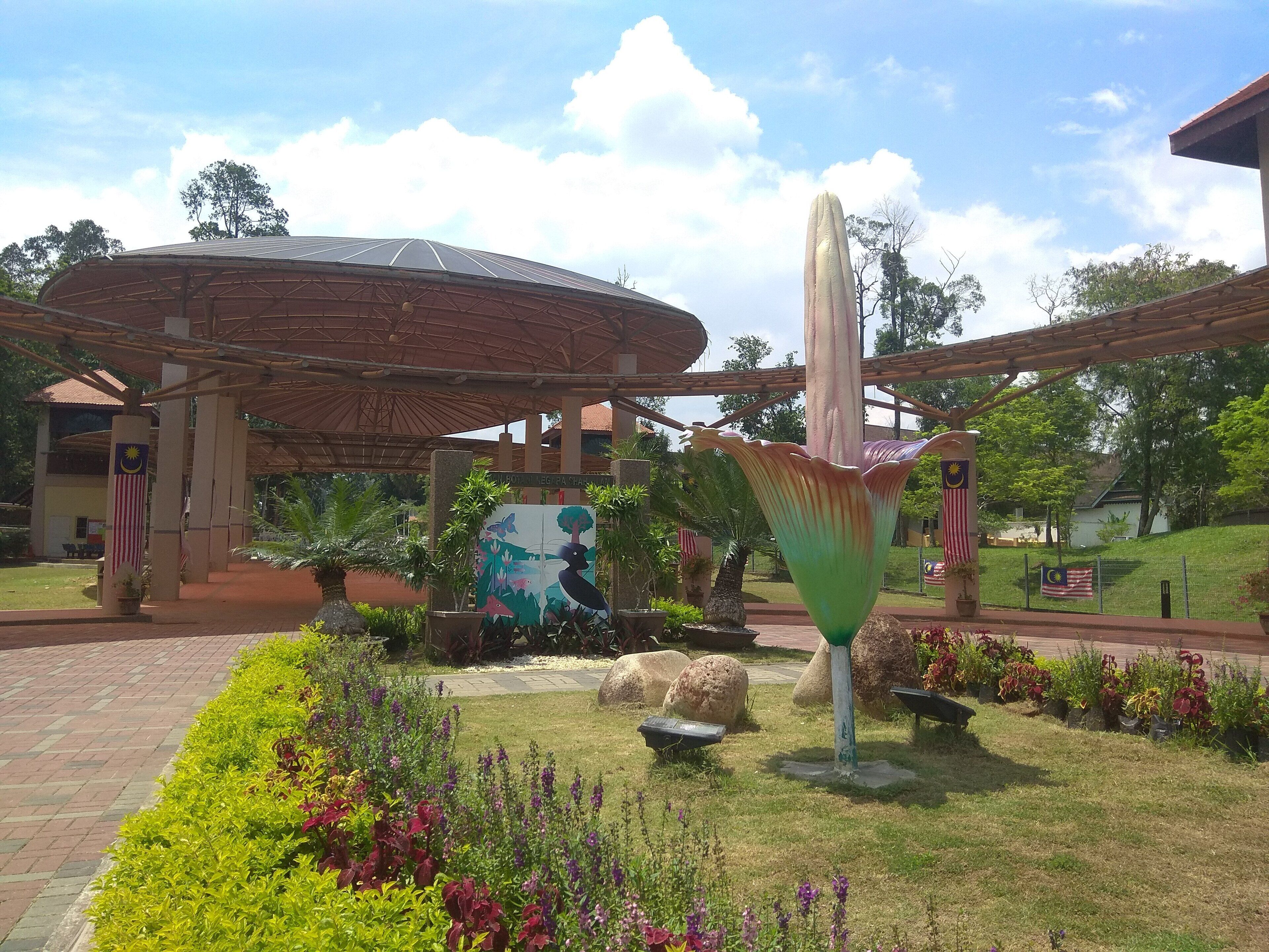 Un jardín con una gran escultura decorativa, un paseo cubierto y un edificio con la bandera de Malasia.