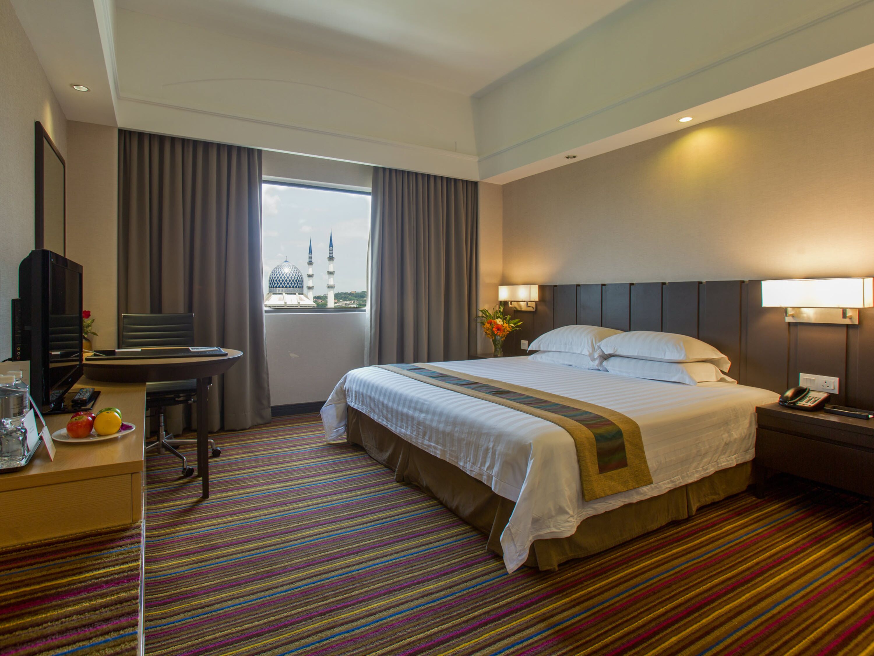 Habitación de hotel con una cama grande, un escritorio con televisor, una silla y vista a una mezquita por la ventana.