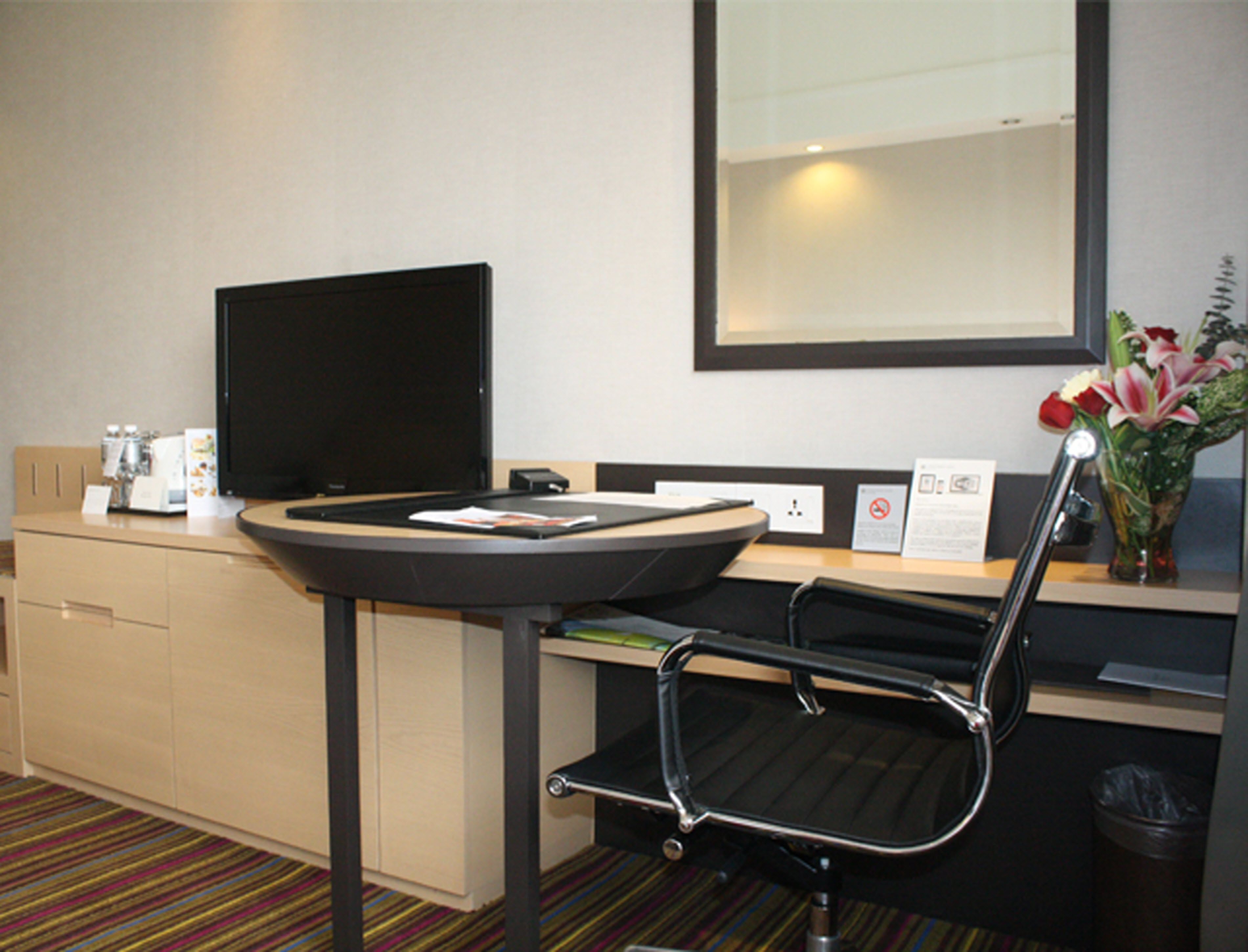 Hotelkamer met een bureau, een stoel, een televisie, een tafeltje, een prullenbak en een vaas met bloemen.