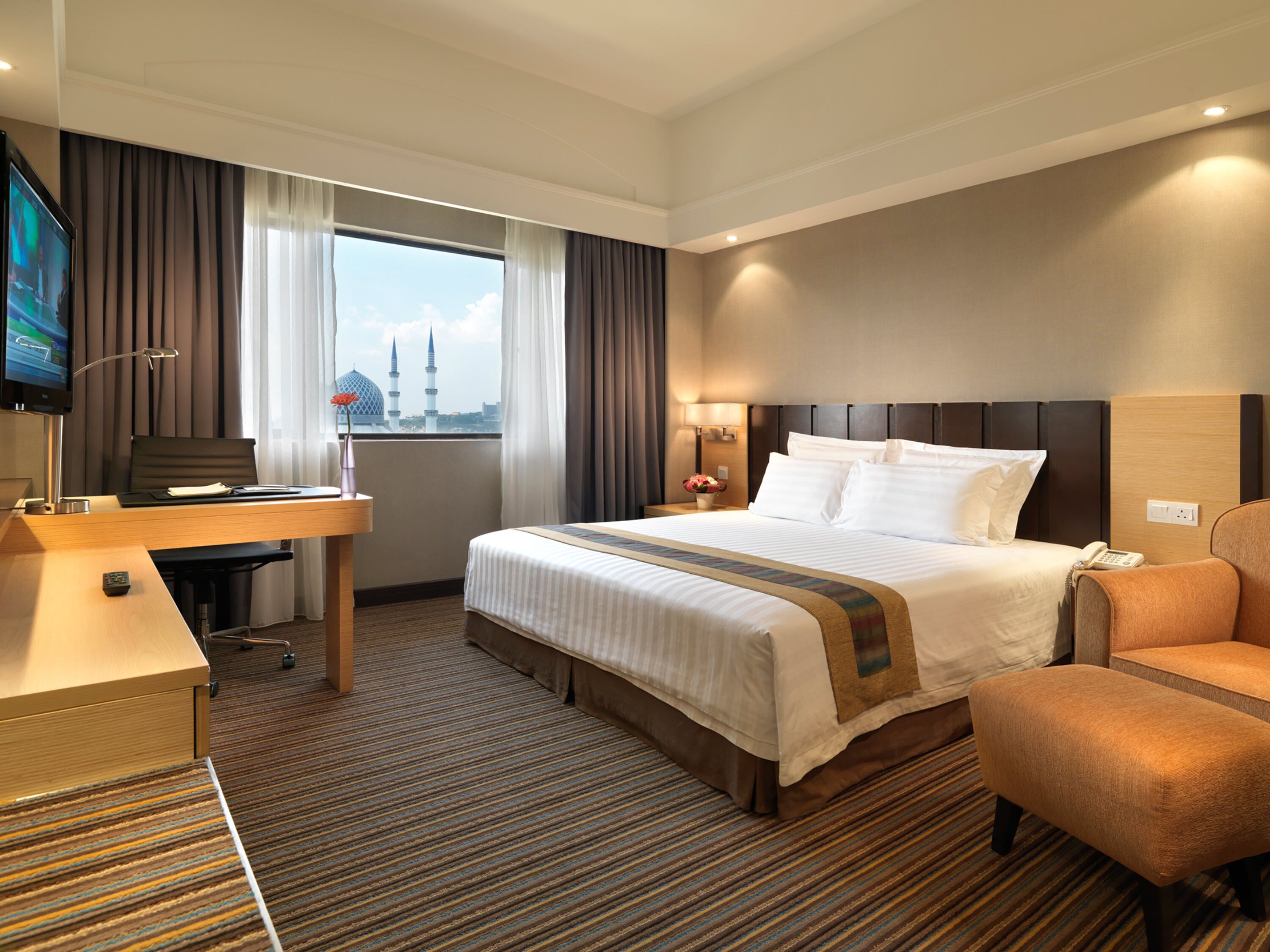 A hotel room with a large bed, a desk, a chair, a TV, and a view of a cityscape.
