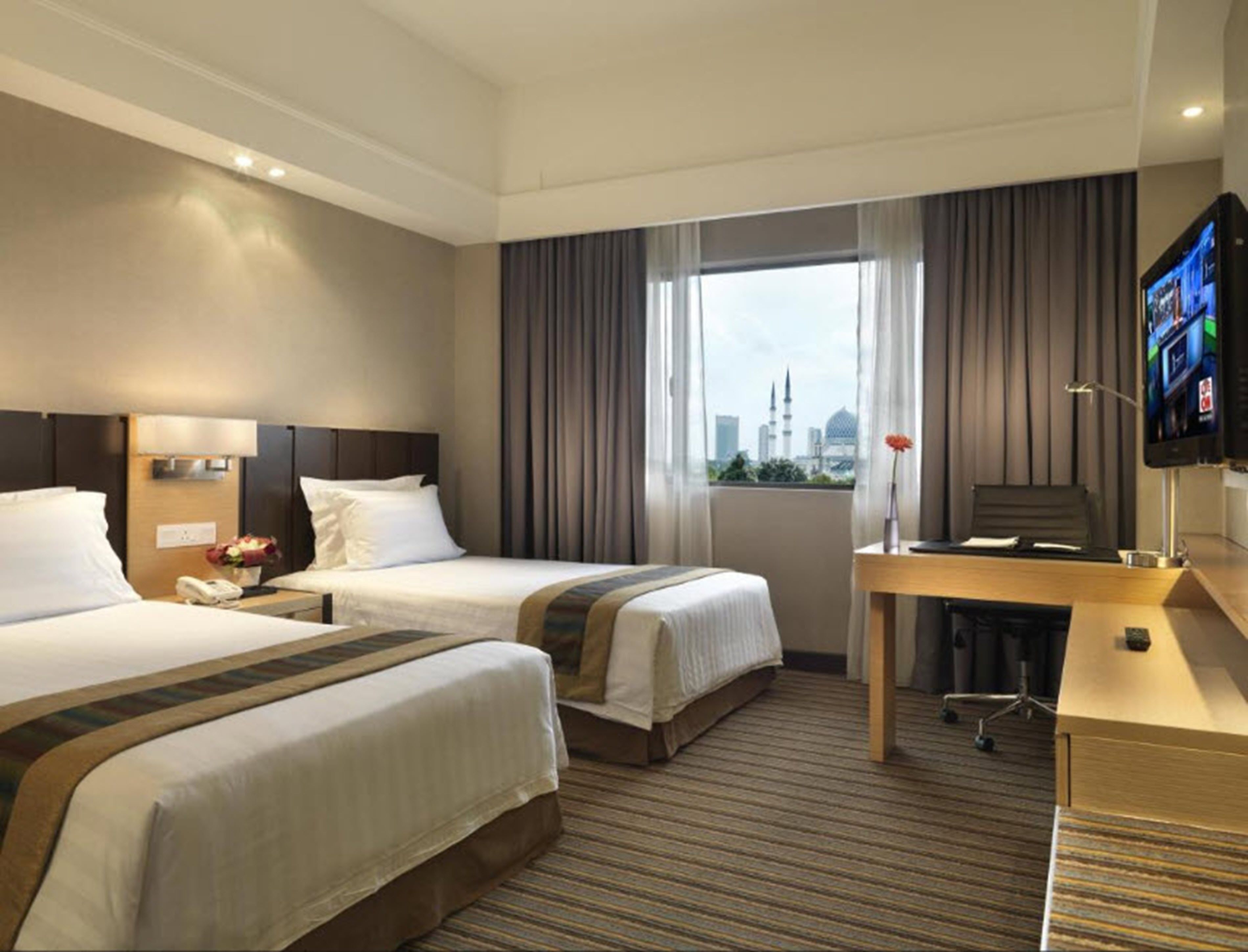 A hotel room with two beds, a desk, a TV, and a view of a cityscape.