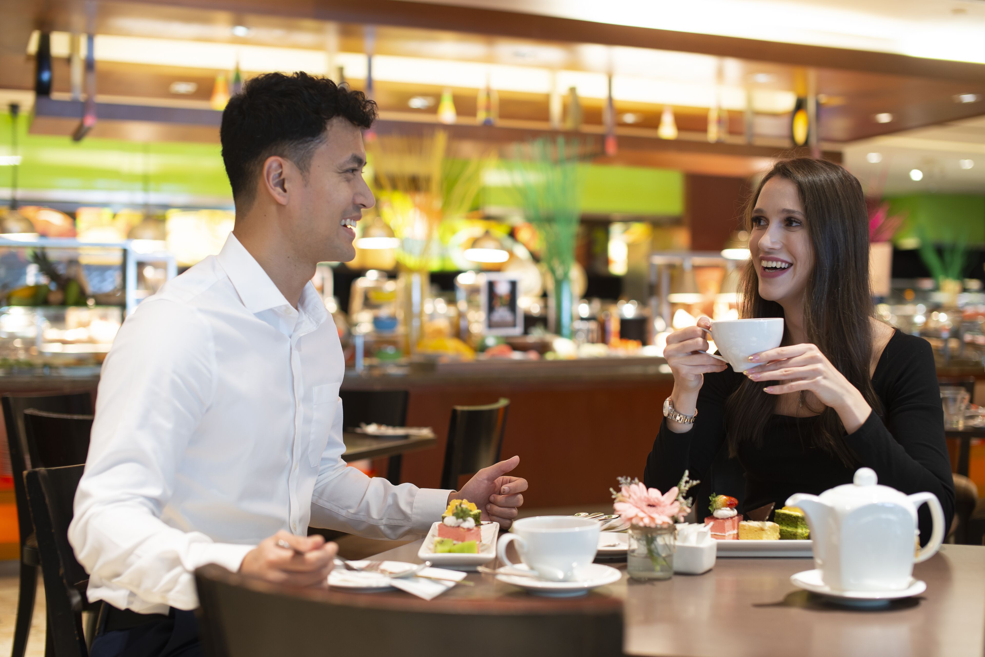 A couple dining in a modern cafe with a variety of desserts and a teapot on the table.