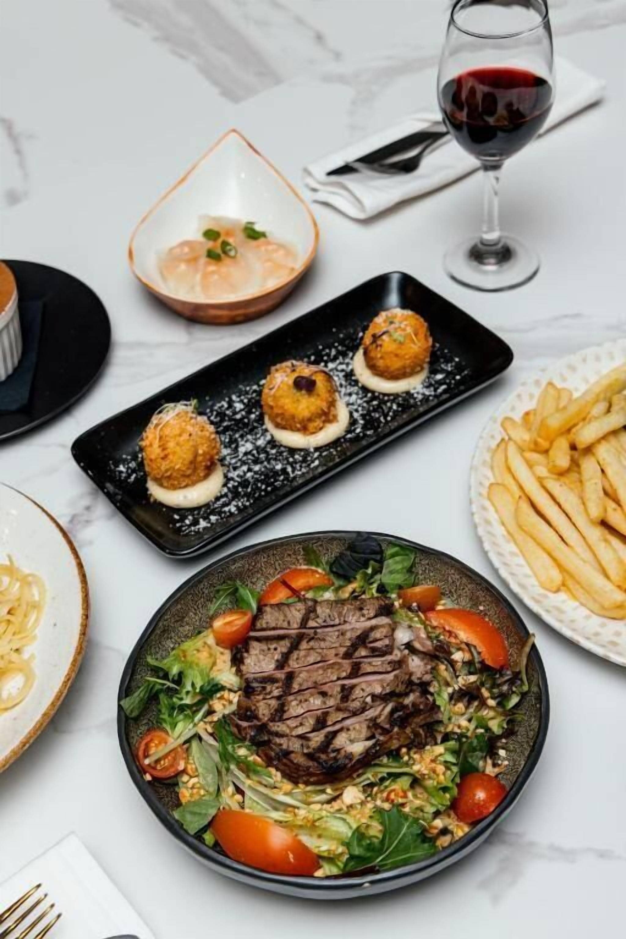 A dining table with various dishes, including a steak with vegetables, a plate of fries, and a glass of red wine.