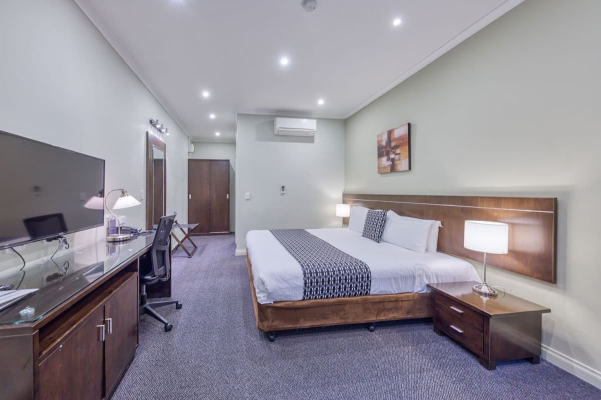 Una habitación de hotel con una cama grande, un escritorio con computadora, una silla, una mesita de noche con lámpara y un equipo de aire acondicionado.
