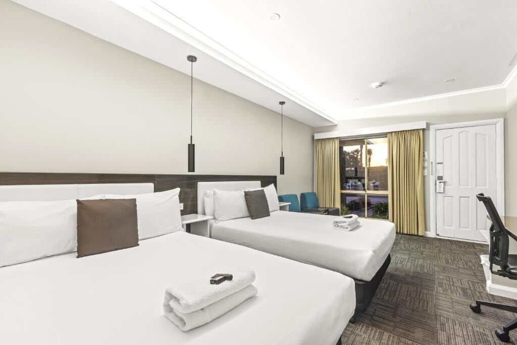 A hotel room with two beds, a desk, a chair, and a view of the outside.