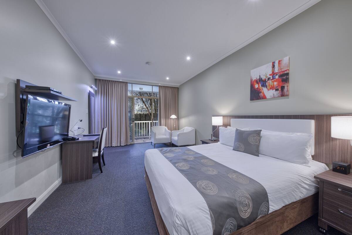 Habitación de hotel con una cama grande, un escritorio con televisor de pantalla plana, dos sillas, una mesita de noche y una ventana con cortinas.