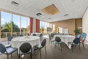 Uma sala de conferências com mesas redondas, cadeiras e uma grande janela com vista para árvores.