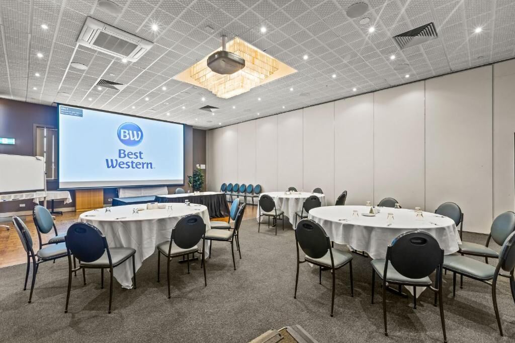 A conference room with round tables and chairs, a projector screen displaying 'BW Best Western,' and a ceiling with recessed lighting.
