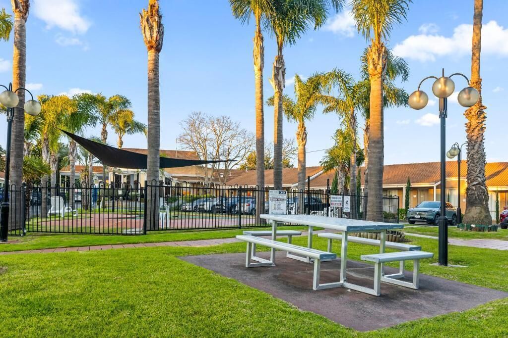A park with picnic tables, a fenced area, palm trees, and a building in the background.