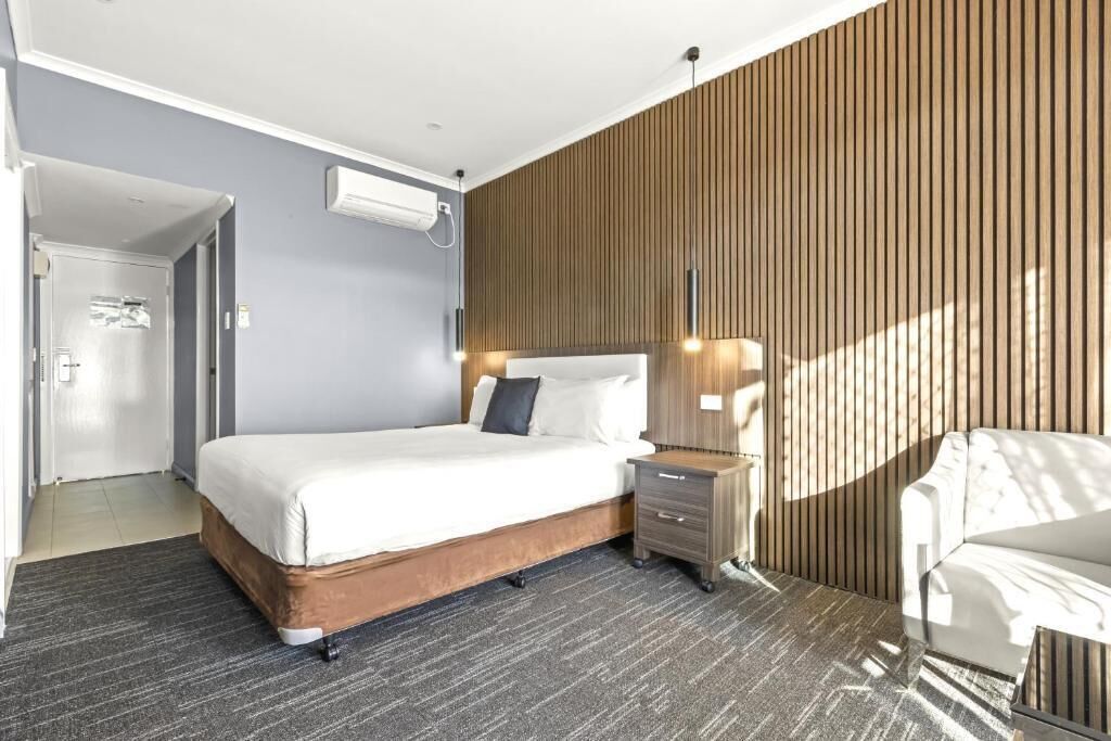 A modern hotel room with a large bed, a bedside table, a chair, and a wooden panel wall.