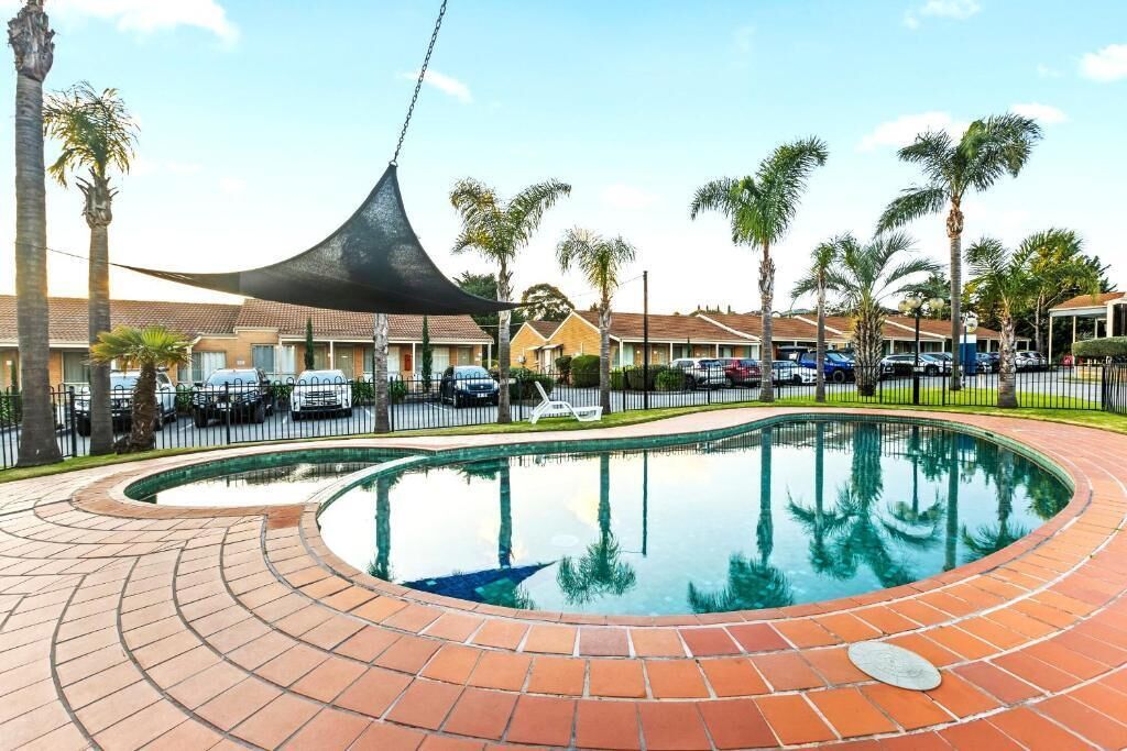 A swimming pool with a curved brick edge, surrounded by palm trees and a row of parked cars.