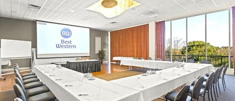 Un salón de conferencias con una larga mesa, sillas, una pantalla de proyector y vistas al exterior.