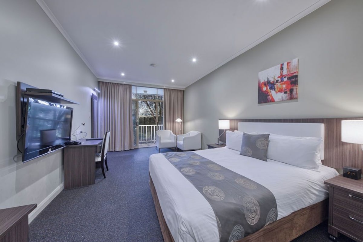 Une chambre d’hôtel comprenant un lit, un bureau avec une télévision, une chaise, une table de chevet et une fenêtre avec des rideaux.