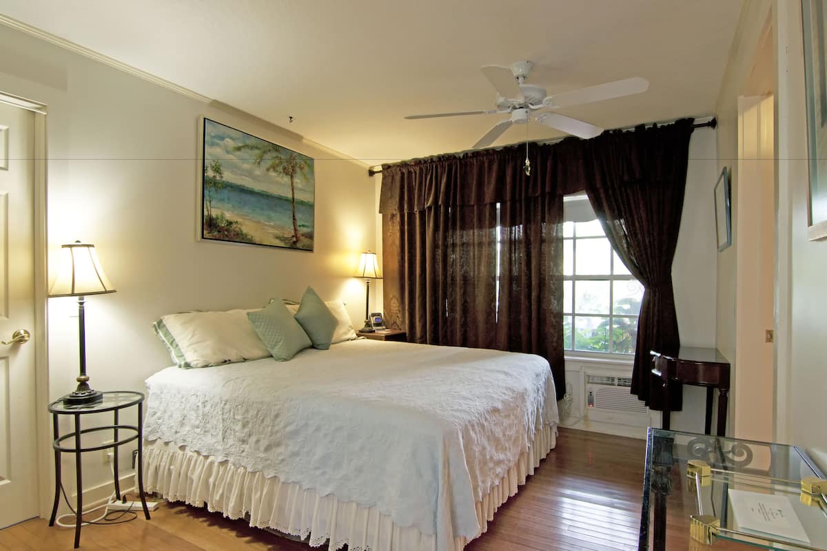 Une chambre à coucher avec un lit, un ventilateur de plafond, une fenêtre avec des rideaux, une table de chevet et une lampe.