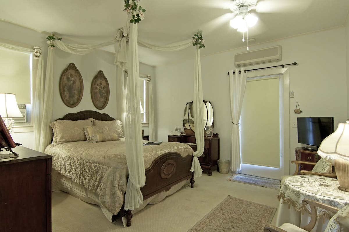Une chambre à coucher avec un lit à baldaquin, une table de chevet, un miroir, une télévision et une petite table avec une lampe.