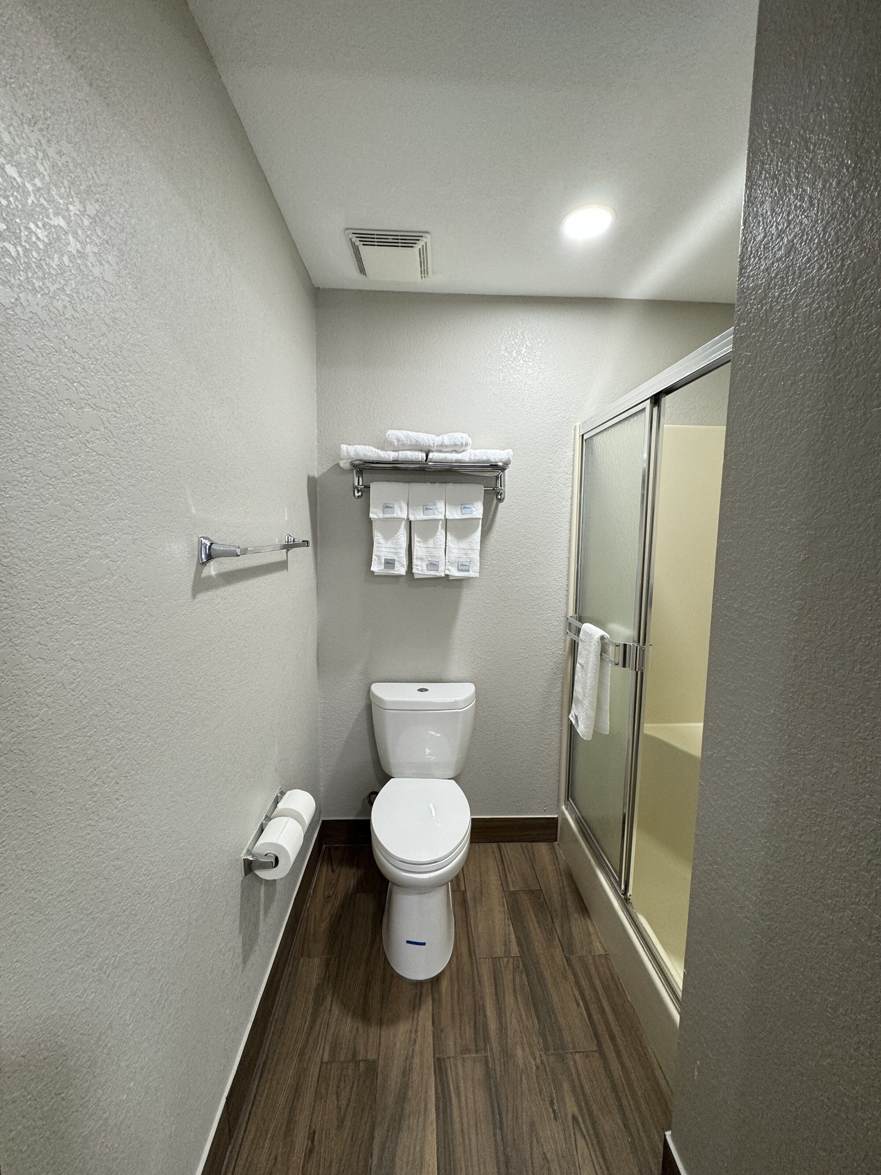 A compact bathroom with a toilet, a towel rack with folded towels, a shower area with a glass door, and a toilet paper holder on the wall.