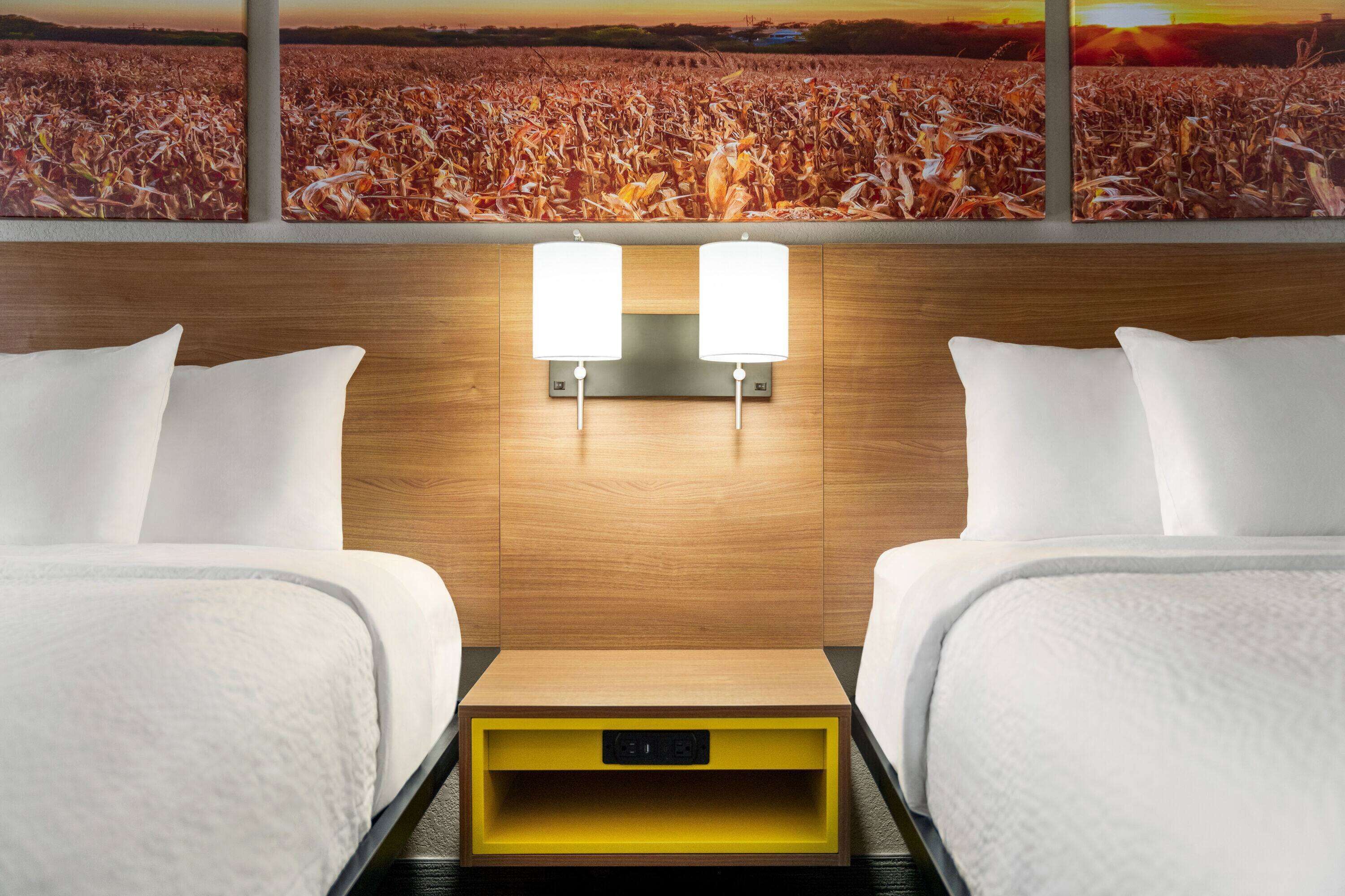 Ein Hotelzimmer mit zwei Betten, einem hölzernen Kopfteil, einem Nachttisch mit Steckdose und einem großen Fenster mit Blick auf ein Feld.