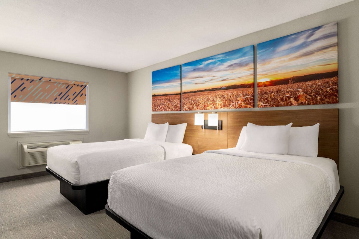 Habitació d'hotel amb dos llits, una finestra gran i una obra d'art penjada a la paret que representa un capvespre sobre un camp.
