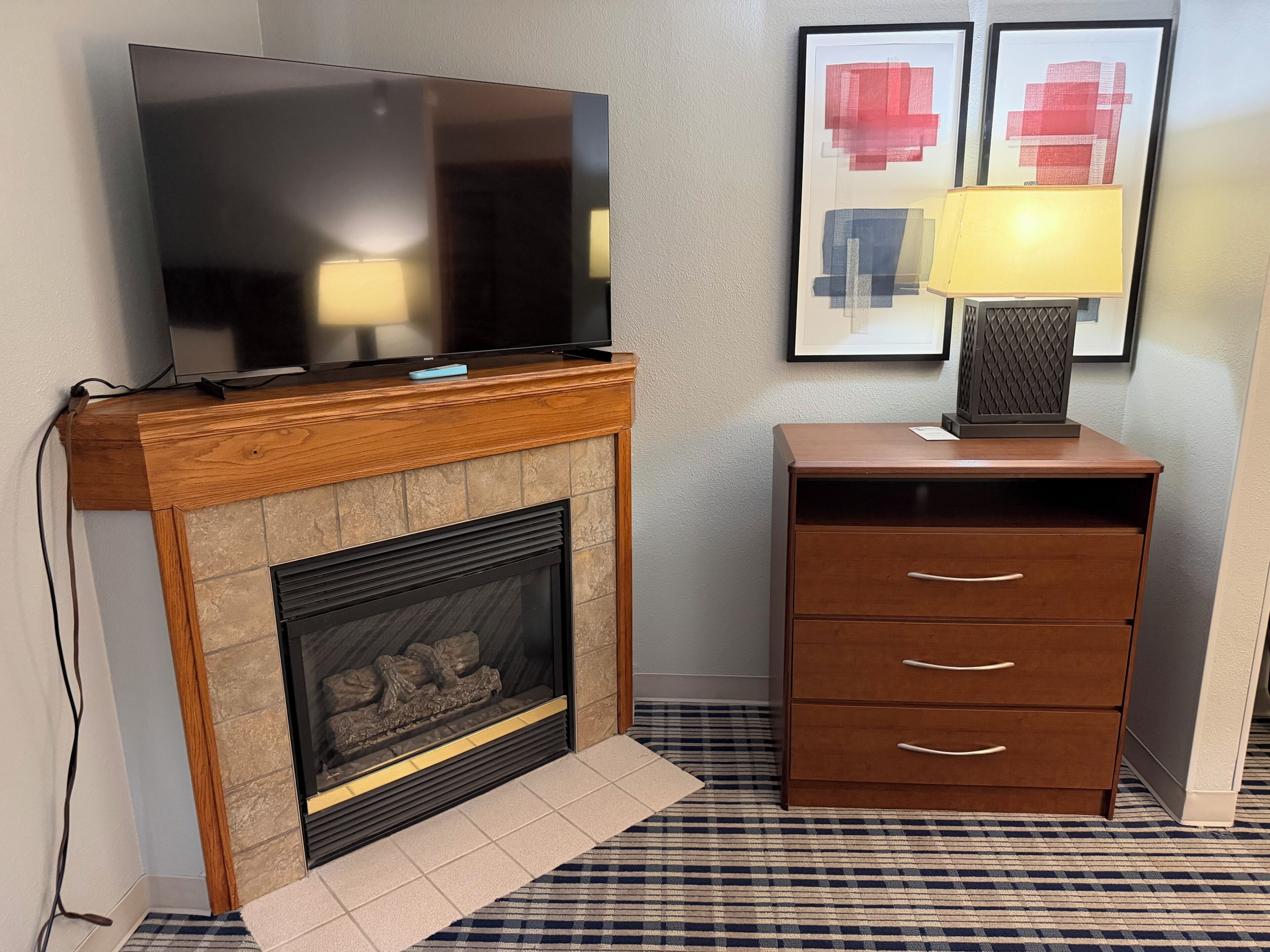 A flat-screen TV on a wooden mantel above a fireplace, a lamp on a wooden cabinet, and two framed abstract paintings on the wall.