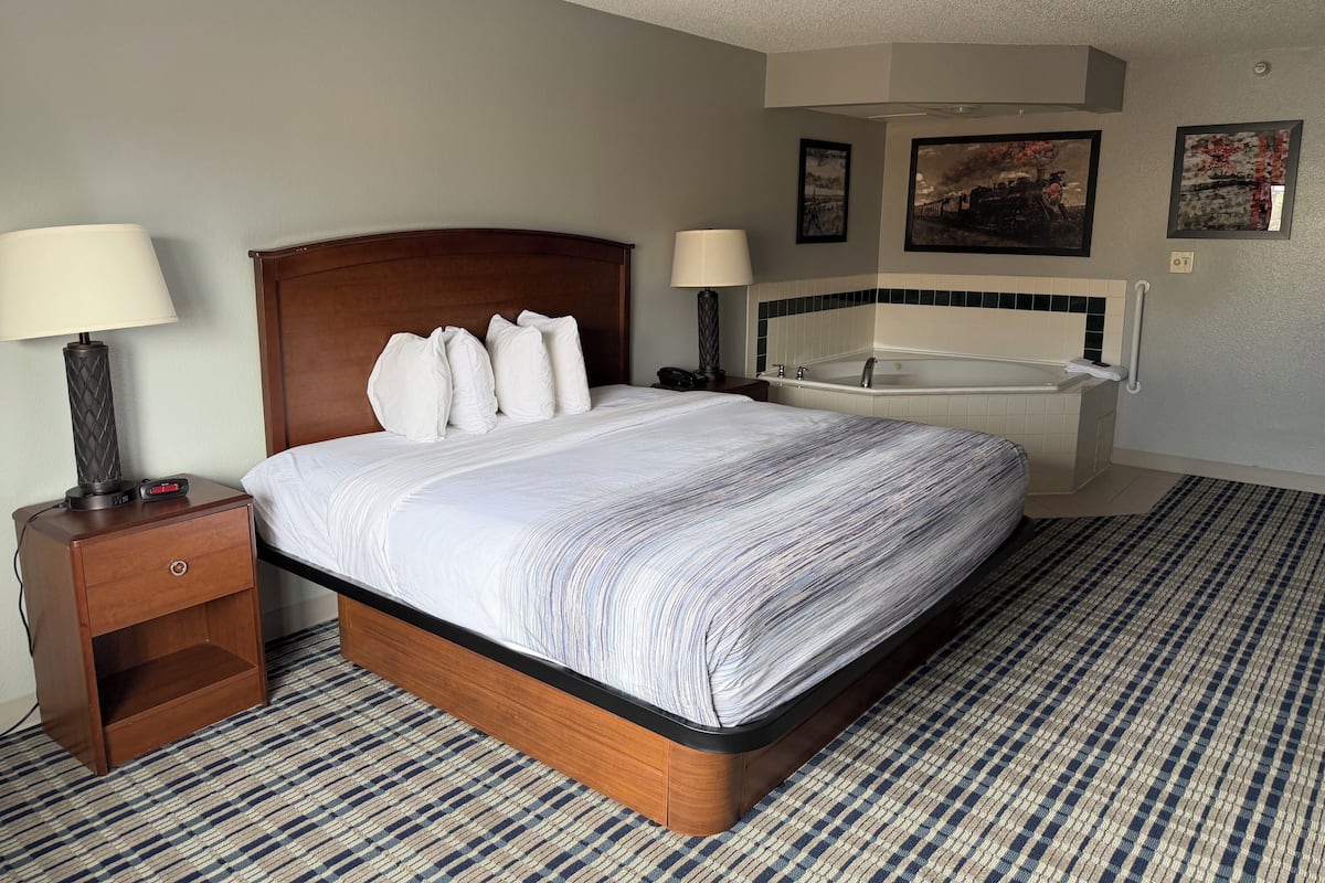 A hotel room with a bed, two bedside lamps, a nightstand, a digital clock, a bedside table, a large window, a bathroom with a bathtub, and a towel rack.
