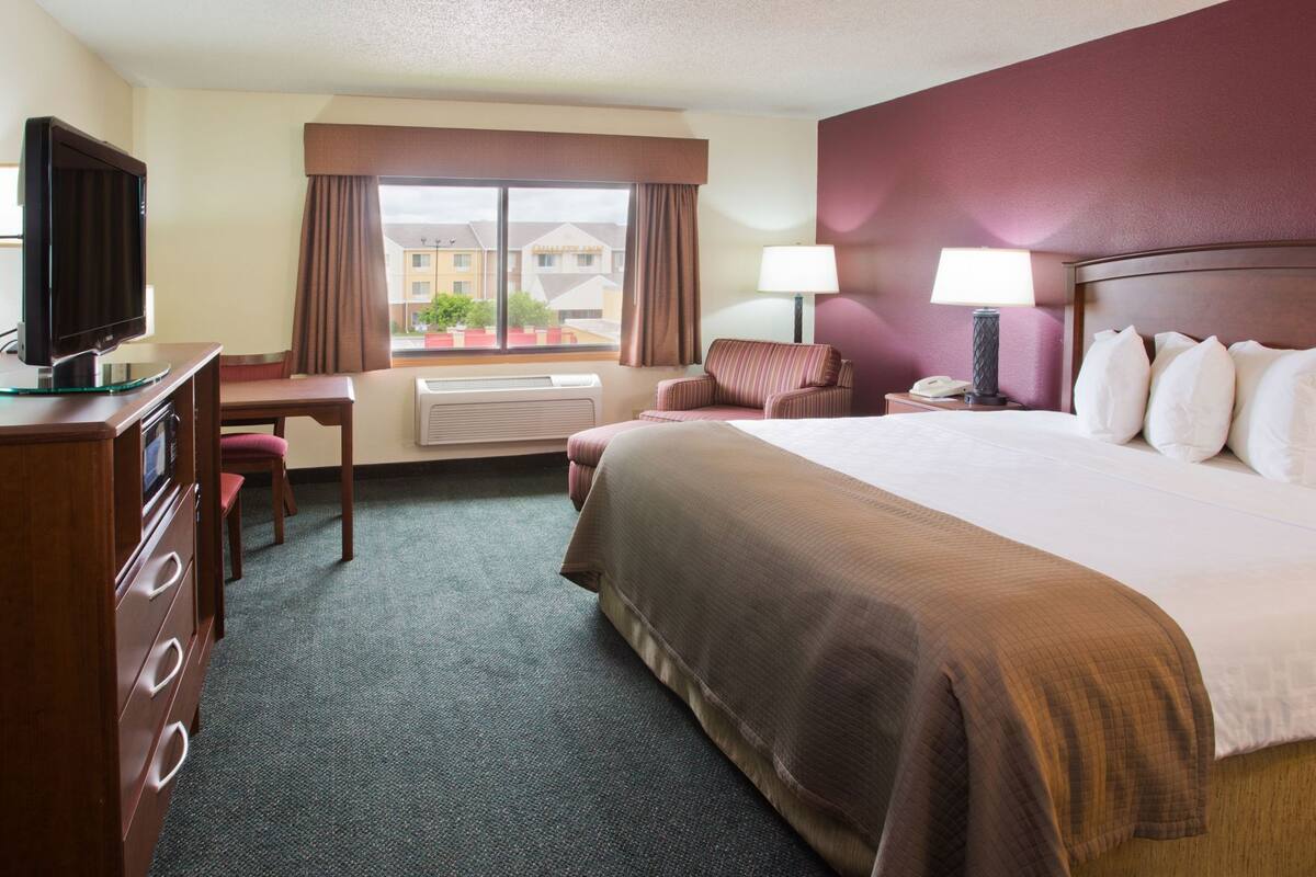 A hotel room with a bed, a desk with a television, a chair, a lamp, and a view of buildings outside the window.